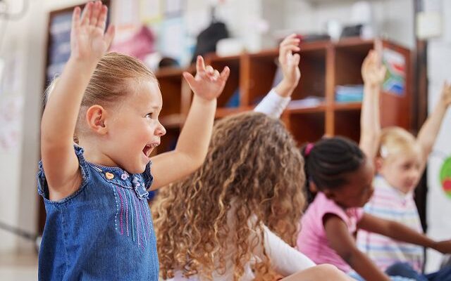 Mehrere Kinder sitzen auf dem Boden und strecken die Hände in die Luft. Im Hintergrund ist ein Regal aus Hold zu sehen.