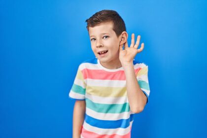 Junge hält eine Hand ans Ohr und lauscht vor blauem Hintergrund.