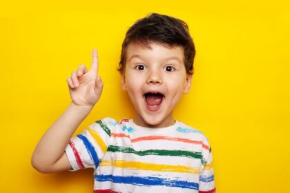 Junge zeigt mit erhobenem Zeigefinger nach oben vor gelbem Hintergrund.