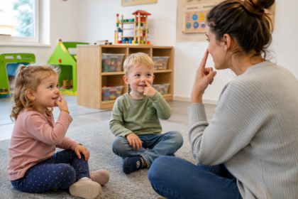 Erzieherin sitzt im Schneidersitz mit zwei Kindern auf dem Boden und alle drei fassen sich mit dem Zeigefinger an die Nase.