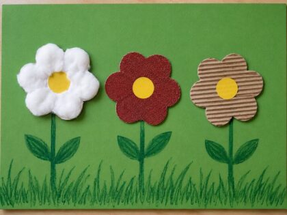 Drei gebastelte Blumen aus Watte, Glitzerpapier und Pappe auf grünem Hintergrund.