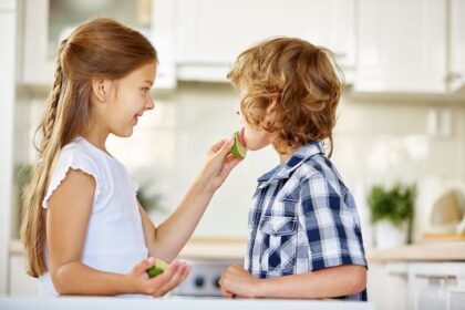 Mädchen gibt einem Jungen in einer Küche ein Gurkenstück zu essen.